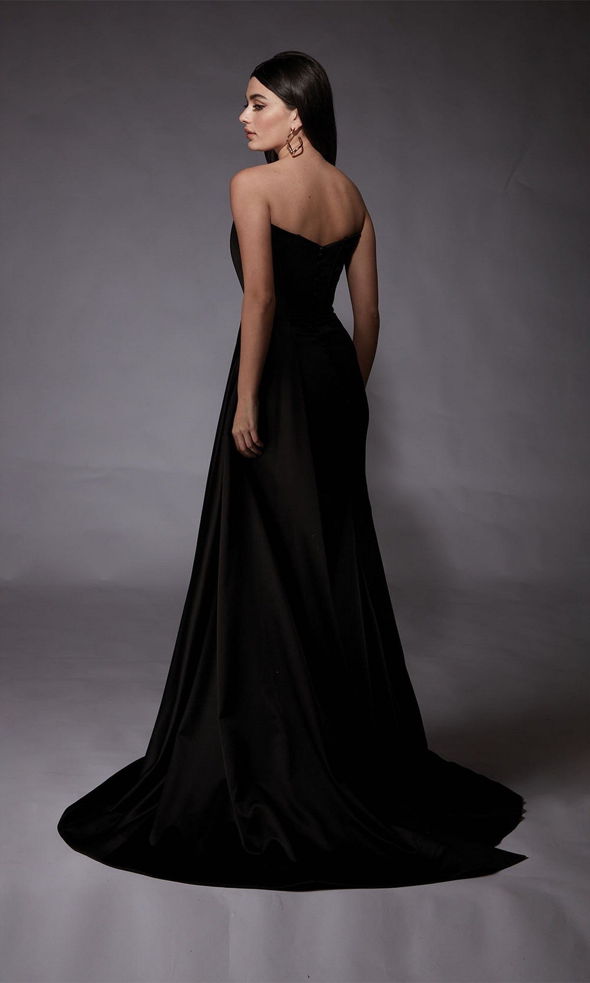 A woman in the Alyce 7093 long formal dress, featuring a strapless satin design and thigh-high slit, stands with her back to the camera against a plain gray backdrop.